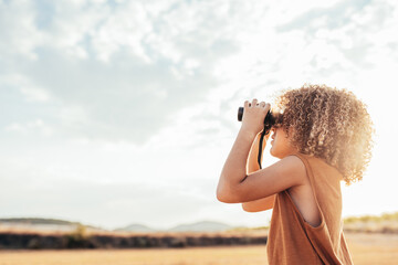 Happy ethnic kid looking through binoculars in field in summer