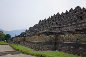 Fototapeta premium Exterior wall of ancient Borobudur temple view from the base of temple with mountain in background and clear blue sky. No people. Popular tourist and Buddhist pilgrimage destination.