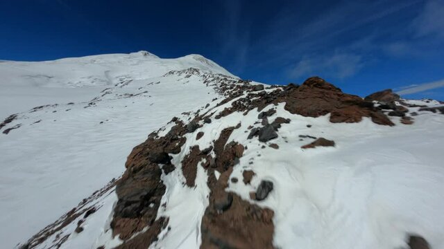 Aerial fpv drone view flying to high mountain peak with skiing trace with ski and snowboard people. Above shot picturesque nature landscape covered by snow track way for climbing and riding at winter