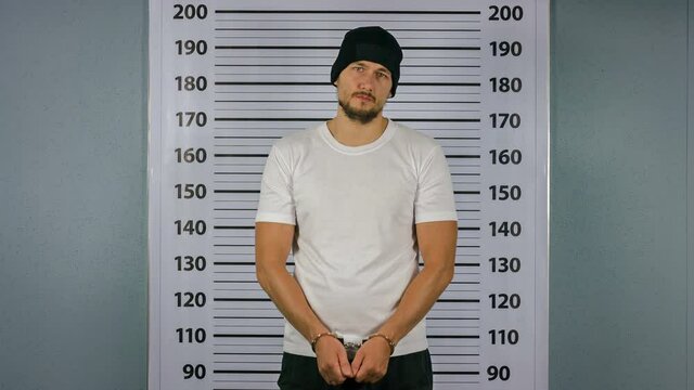 Arrested a man in handcuffs standing near a white board with a scale. Mugshot