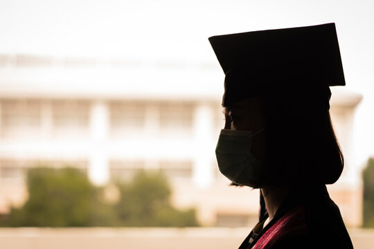 Silhouette photo of a young happy Asian woman university graduate in graduation gown and cap or mortarboard wears face mask to celebrate graduation day