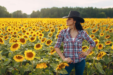 Young woman agronomist wearing cowboy hat, plaid shirt and jeans on sunflower field. Summer harvest...