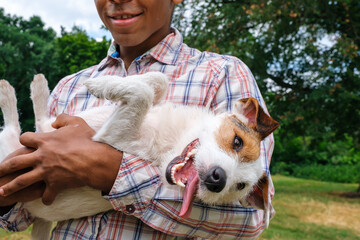Close-up Brazilian man holding a funny Jack Russell terrier in his arms in summer outdoors. The puppy smiles cheerfully and shows tongue. Pet care