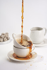 pouring coffee into a white porcelain cup with cappuccino stacked in another mug on a white saucer plate on a white surface
