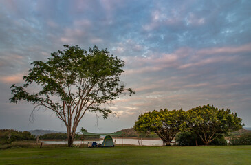 Fototapeta premium View of a camping tent with awning pitched On the grass under beautiful big trees with branches at the campsite lake shore with mountain range in background. Selective focus.