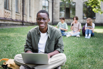 Cheerful african american student using laptop on lawn outdoors