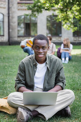 Obraz premium Smiling african american student using laptop on lawn