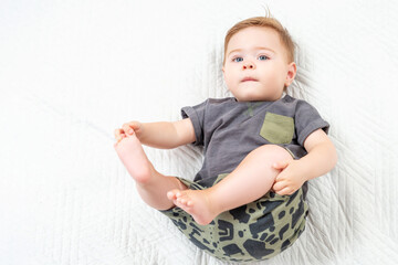 Fototapeta premium Baby boy in a khaki suit lying on white bed and holding legs. Portrait of adorable toddler on white background, copy space