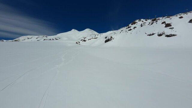Aerial fpv drone view fast flight over snowy mountain Elbrus peak cinematic scenery landscape with skiing track. Above shot natural winter scenery cliff tops surrounded by clear blue sky sunny day