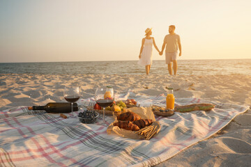 Couple walking by sea, focus on blanket with food and snacks. Summer picnic