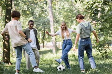Fototapeta premium Excited girl playing football with multiethnic teenagers in park