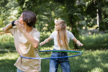 Side view of smiling girl twisting hula hoop near blurred friend in park