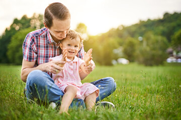 Naklejka na ściany i meble Young daddy sitting in a park in the grass with his little toddler daughter on his lap - 20s husband plays with his child and teaches to clap her - Parenthood and childhood family lifestyle concept
