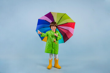 a little boy in a green raincoat and yellow rubber boots stands with a multicolored umbrella stands on a blue background with a place for text