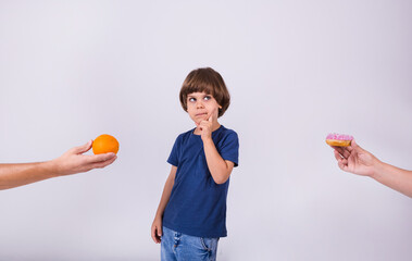 a thoughtful little boy in a T-shirt makes a choice between an orange and a donut on a white background with a place for text