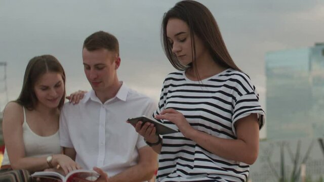 Two girls and a guy sitting in the park during the day reading an e-book