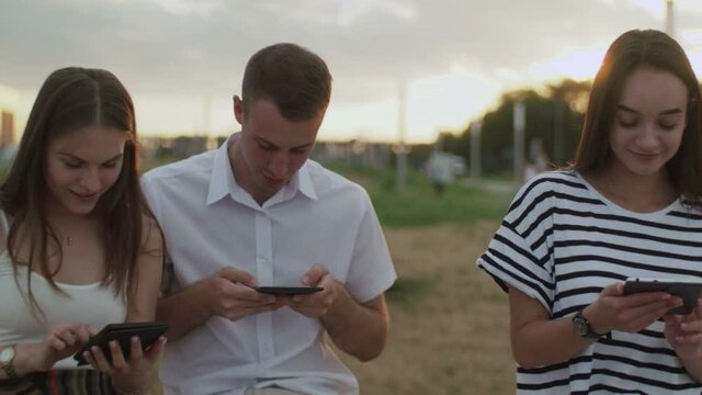 Two girls and a guy sitting in the park during the day reading an e-book