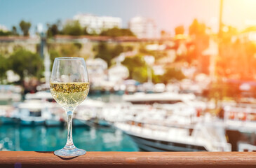 Glass of white wine against the backdrop of the Mediterranean sea and the port with yachts in a tourist town in the summer under sunlight. Summer, travel, lifestyle, relaxation, and enjoyment concept.