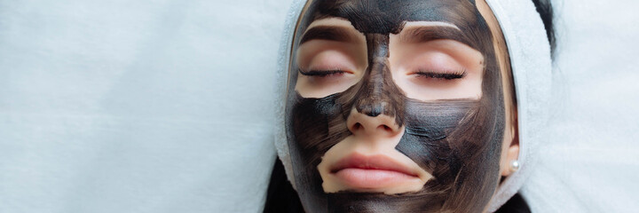 banner . cosmetologist applying black mask on pretty woman face wearing black gloves, gorgeous woman in spa having facial procedures