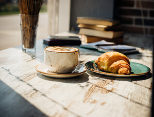 Cappuccino and croissant are lying on the table in a cafe against the background of a window and a stack of books.