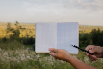 An open notebook in a cell on nature close-up. Hands point with a pen to an unfolded notebook. A...