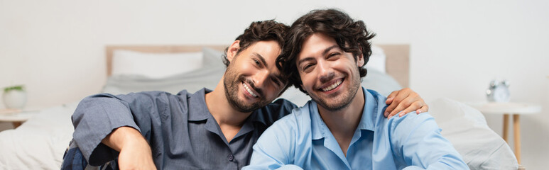 positive young gay couple hugging in bedroom, banner