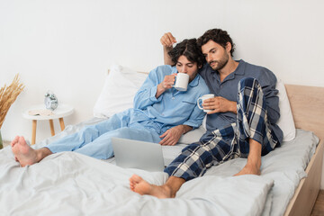 gay couple holding cups of tea and watching movie on laptop in bed