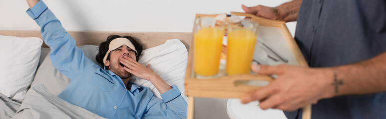 man in eye mask yawning near blurred boyfriend holding tray with orange juice, banner