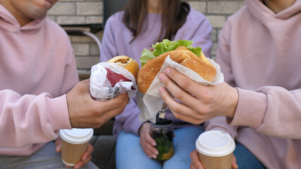 Group of young friends eating takeaway street food while sitting on a bench and chewing on vegan hamburgers. Three happy friends eating healthy food in the park