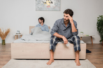 man sitting on bed and looking aside after disagreement with boyfriend on blurred background,...