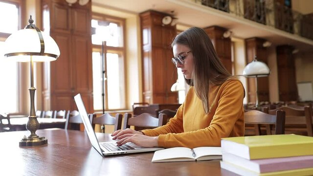 A female student in glasses and a sweater is studying at the table in the library, using a laptop and books. Girl preparing for exams - student lifestyle.