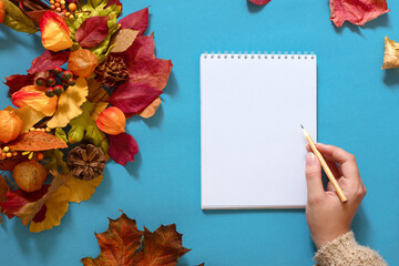 autumn layout with wreath of fallen leaves and blank white sheet for text, hand with pencil, mock...