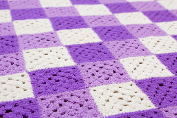 Woolen, knitted blanket. Consists of multi-colored squares knitted with woolen threads. The squares are purple, purple, white and sewn together. Angle view. The background is out of focus.