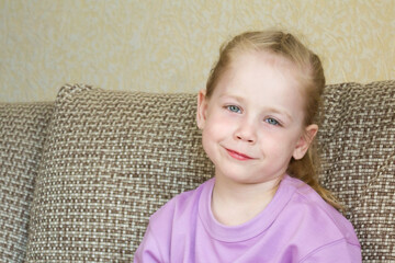 Portrait of a blonde girl with braided hair in a purple t-shirt, leaning back on sofa cushions and looking at the camera