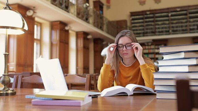 A tired, exhausted student is sitting in the library at a table with books and a laptop, preparing for the exam, taking a break, taking off her glasses and stretching her neck.