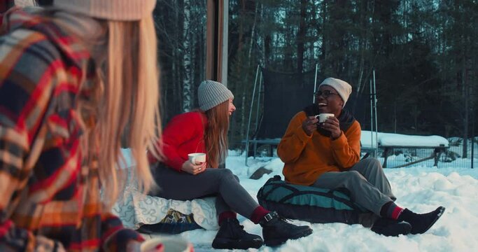 Cheerful happy multiethnic friends drink tea, talk smiling sitting on beanbags at cozy winter house terrace slow motion.