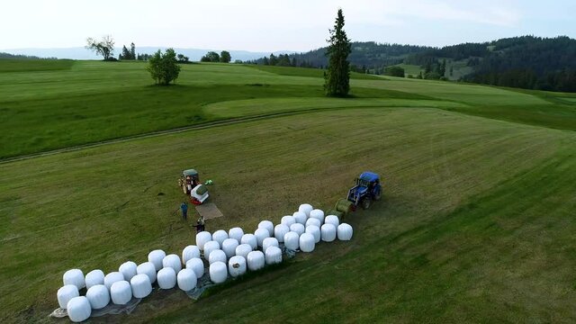 Aerial view of haymaking. Farmers working in the field. Agricultural baler machine and tractor. Fields in the mountain village. High mountain range in the background. 