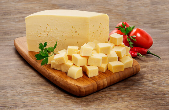 Queijo Muzzarela em pe&ccedil;a e em cubos, sobre t&aacute;bua de madeira em fundo de madeira r&uacute;stica.