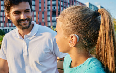 Positive female child with a hearing impairment uses a hearing aid to communicate with her father outdoor. Hearing solutions