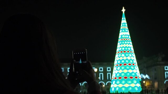 A small christmas outdoortree decorated with lights and a woman taking picture of it with her phone