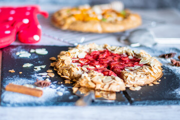 Homemade galette pie cake with strawberries and almond, placed on black desk and wooden table. Red towel with cooking tools on the side