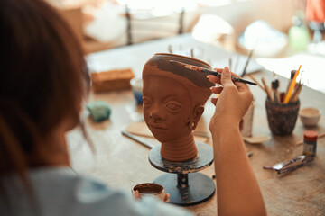 Young woman ceramist painting on clay sculpture in workshop