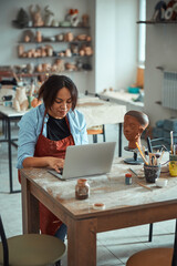 Fototapeta premium Charming female ceramist using laptop in pottery workshop