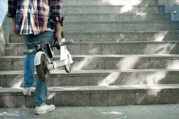 Close-up of male hands holding an electric scooter near the stairs. Faceless guy climb up the stairs. Ecological technological lifestyle. © Andrii Lysenko