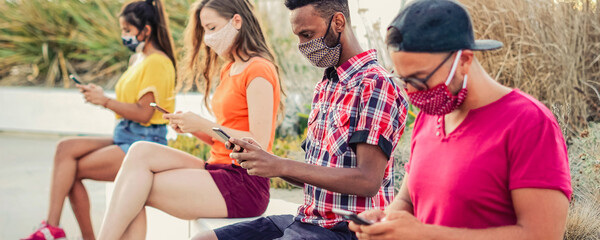 Friends using their smartphones in covid 19 times protected with face mask - Young people using mobile device in distance outdoors - Powered by Adobe