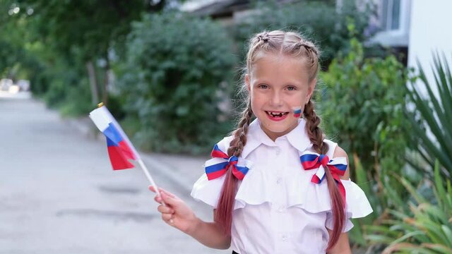 A schoolgirl stands in the street and waving the Russian flag. Happy little girl with face painting of Russian symbols. Day of the Russian flag.