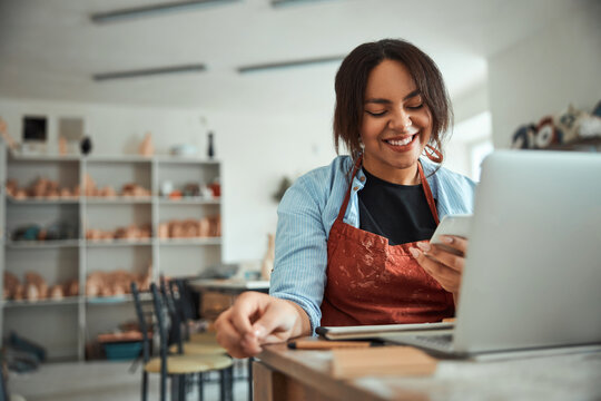 Cheerful woman using mobile phone and laptop in pottery workshop
