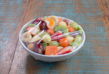 Salada de frutas em tijela coberta com fime plástico no fundo de mesa rústico.