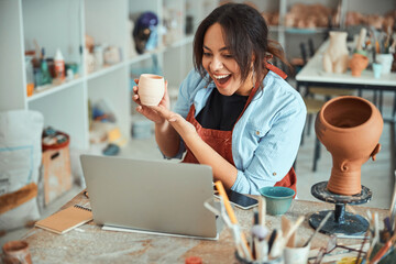 Joyful female potter holding clay pot and using laptop in workshop