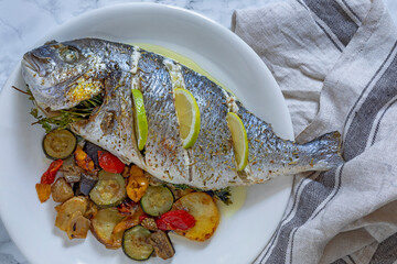 Plate with a whole sea bream with lemon slices and mixed grilled vegetables.